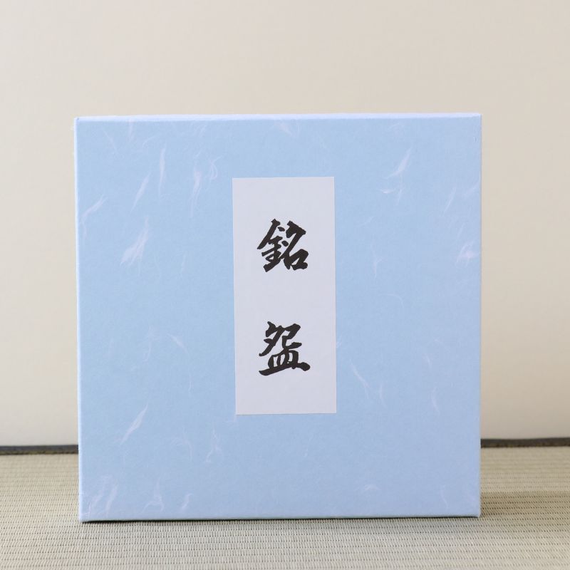 A square light blue box with a white vertical label featuring black Japanese characters, photographed against a plain beige wall and textured surface.