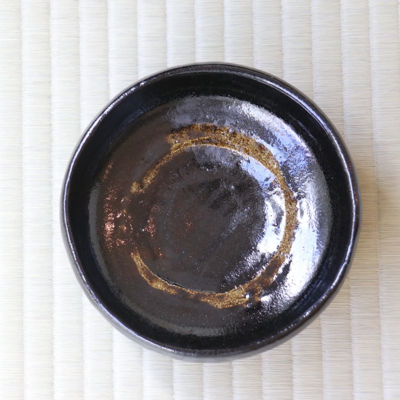 A round black ceramic bowl with a glossy finish and a decorative gold-brown ring near the rim, photographed from directly above on a light beige textured mat.