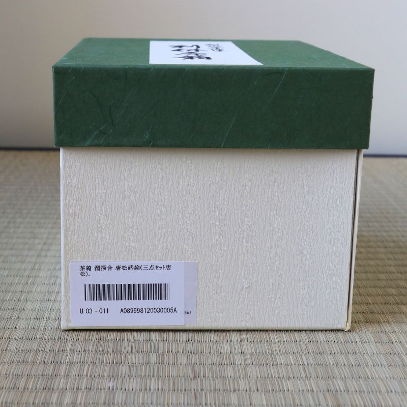 A square product box with a dark green lid and a cream-colored body, placed on a woven mat. A white label with Japanese characters is visible on the lid, and a barcode label is attached to the side.