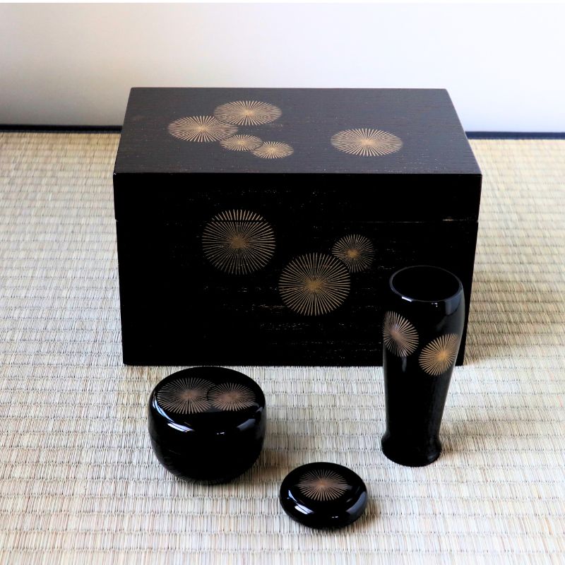 A black lacquerware set with gold sunburst patterns, including a rectangular box, a tall vase, a round lidded container, and a small dish, arranged on a woven mat.