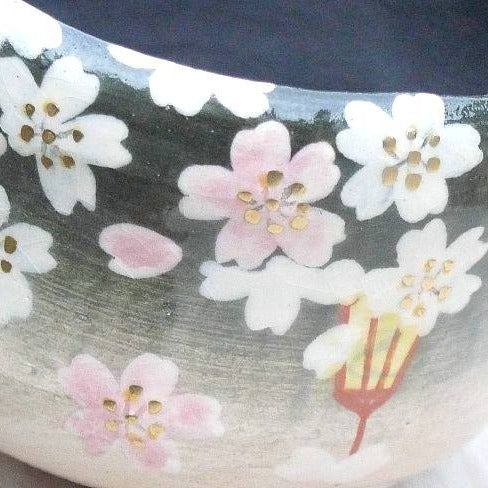 Close-up of a ceramic bowl featuring hand-painted cherry blossoms in white and pink with gold centers on a dark and light gray gradient background.
