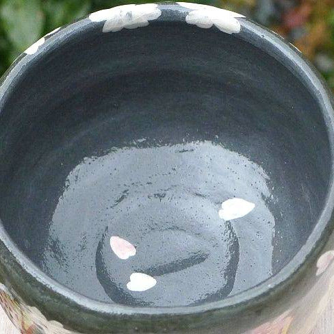 A close-up of a dark glazed ceramic bowl with white and pink floral patterns around the rim, set outdoors on a light-colored surface with blurred green foliage in the background.
