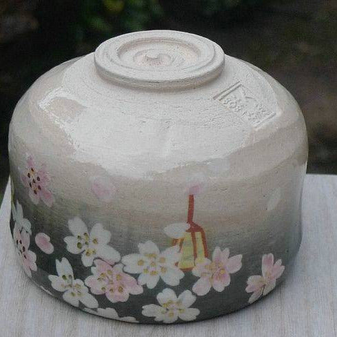 A handmade ceramic bowl with a glossy glaze, featuring a painted cherry blossom pattern in white and pink on a dark gray base, with a small red and yellow lantern illustration. The bowl is photographed outdoors on a light-colored wooden surface with a blurred green garden background.