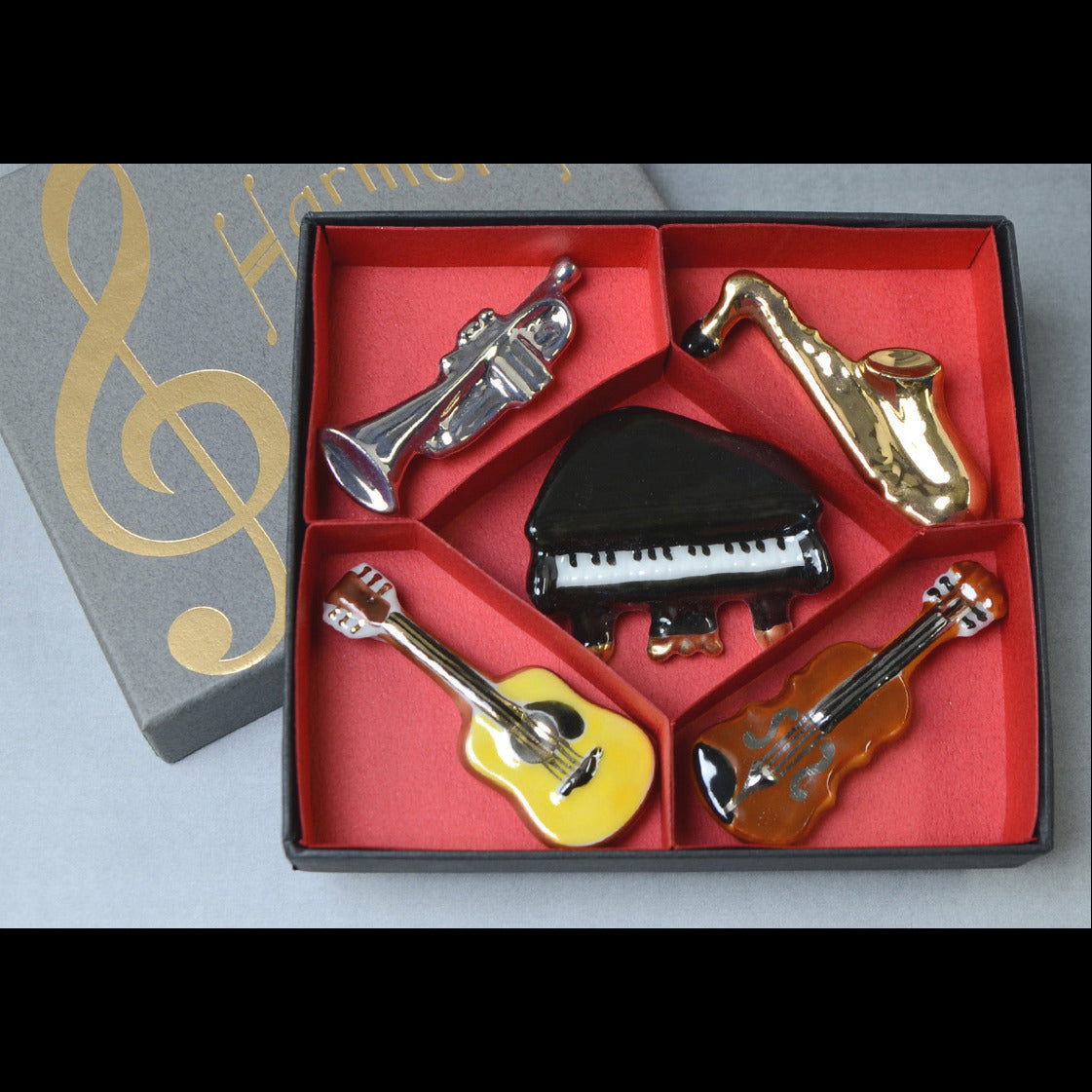 A set of miniature metal music instrument charms, including a trumpet, saxophone, piano, guitar, and violin, displayed in a red-lined compartmentalized box next to its gray gift box with gold lettering.