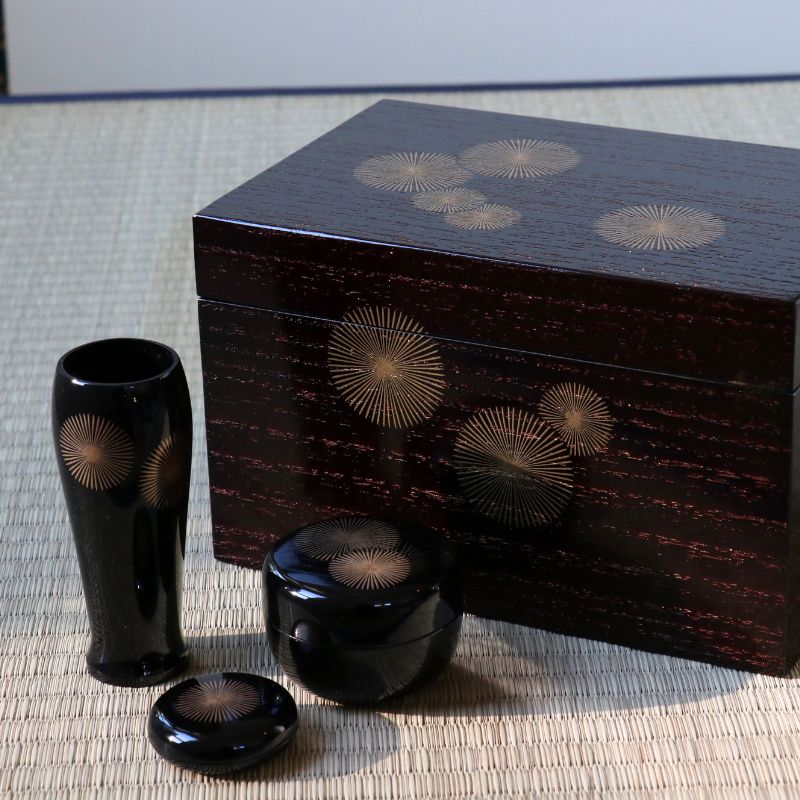 A set of glossy black lacquered Japanese decorative objects, including a tall vase, a small round jar, a lid, and a large rectangular storage box, all decorated with gold-toned sunburst patterns on a woven mat background.