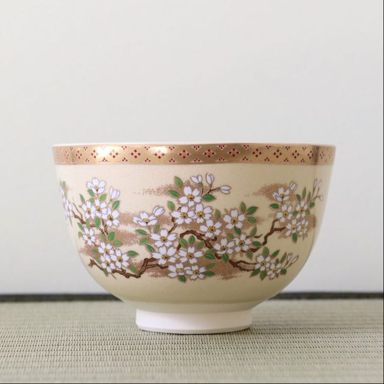 A ceramic bowl with a cream-colored glaze, decorated with a hand-painted cherry blossom branch pattern in white, green, and brown, featuring a gold patterned rim.