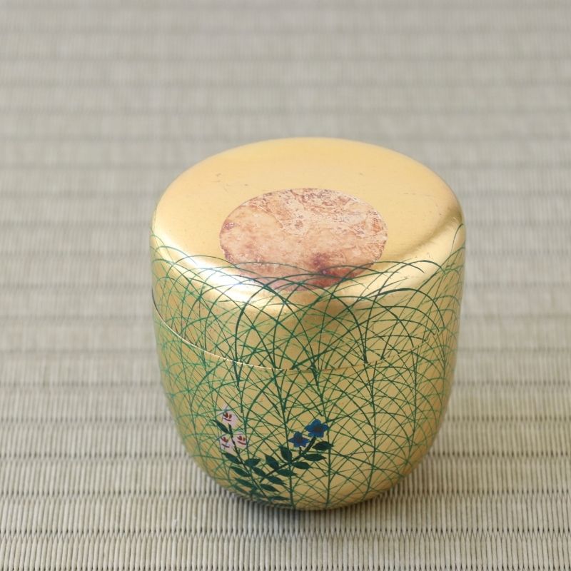 A small cylindrical ceramic container with a pale yellow body, decorated with a green geometric lattice pattern and delicate blue and pink floral accents near the base. The lid is a matching pale yellow with a circular textured brown patch in the center.