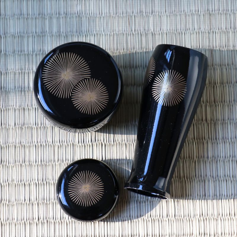 Three-piece glossy black lacquer set with metallic gold starburst patterns, including a tall vase, a round container, and a small lid, arranged on a woven straw mat.