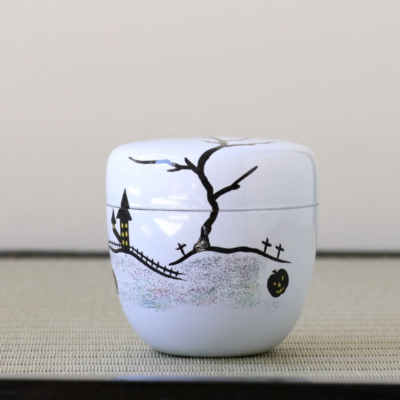 A small white ceramic lidded container decorated with a black hand-drawn style Halloween scene, including a bare tree, haunted house, fence, crosses, and a jack-o'-lantern, shown on a textured beige surface against a plain light background.