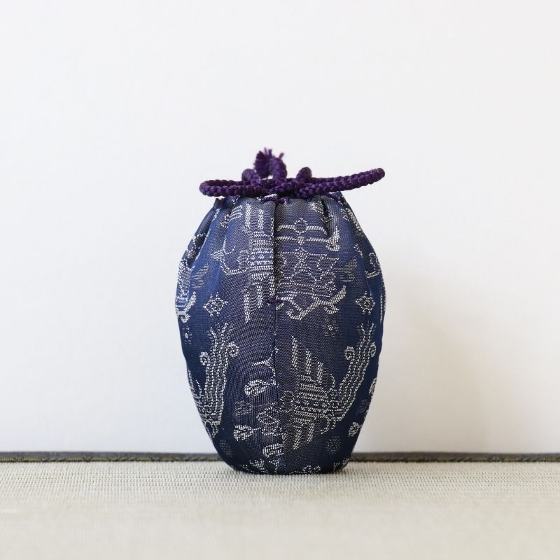 A small drawstring pouch made of dark navy blue patterned fabric with a purple cord tie, standing upright on a light textured surface against a plain off-white background.