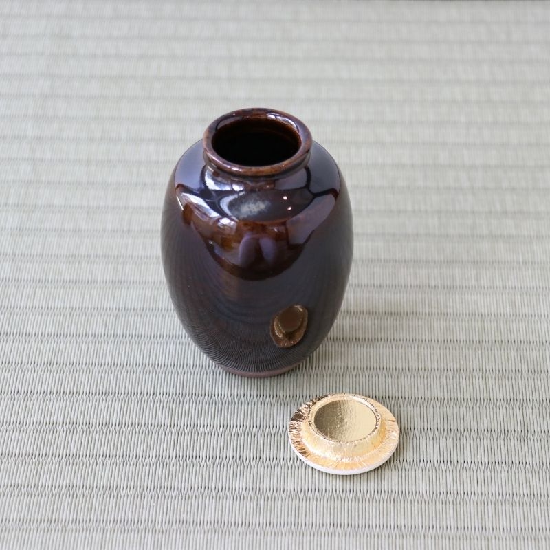 A small glossy dark brown ceramic jar with a matching lid placed beside it on a light gray woven mat background.