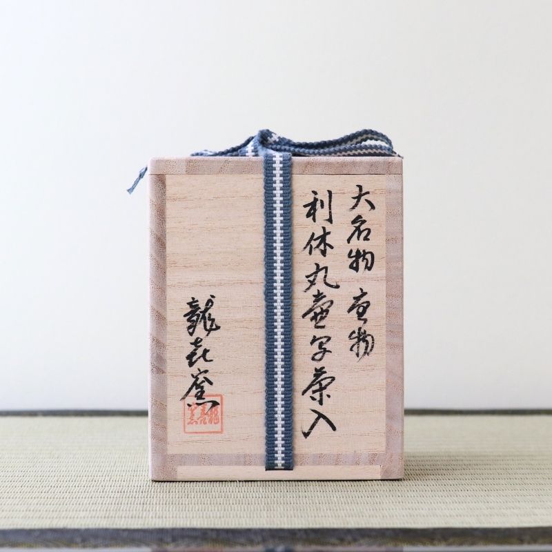 A light natural wood Japanese box with black calligraphy and a blue-and-white striped cord, photographed against a plain light background.