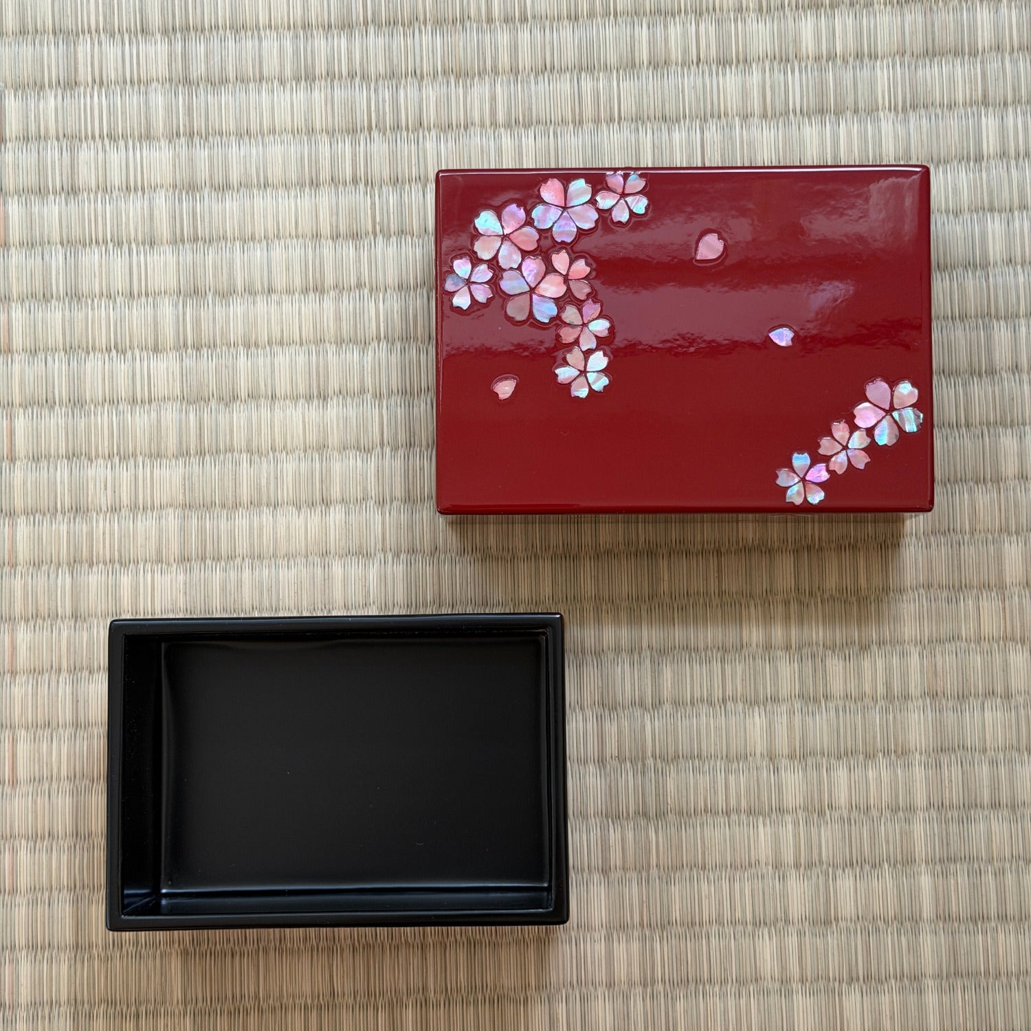 Red lacquerware box with floral design next to a black lacquerware box on a woven mat.