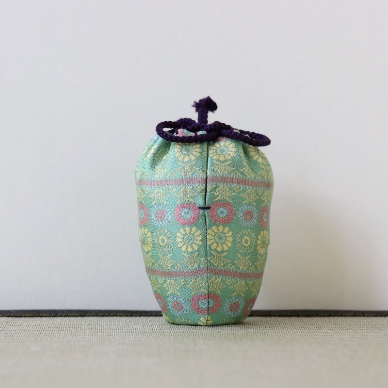 A small fabric drawstring pouch with a light green base, decorated with a colorful floral pattern in pink, yellow, and blue, finished with a dark purple knotted cord closure, set against a plain light gray background.