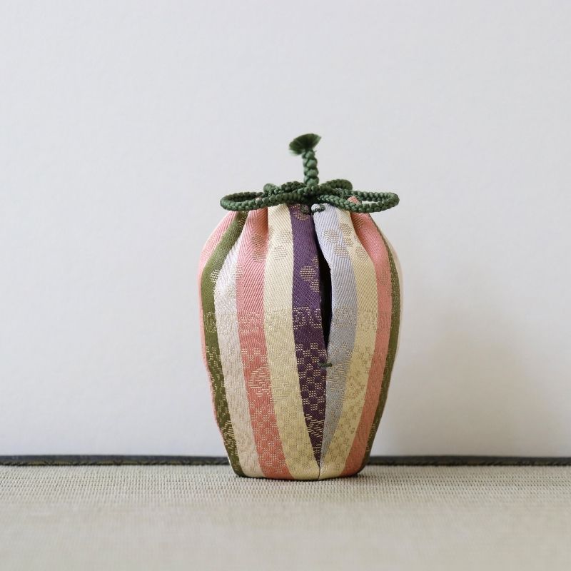 A small decorative fabric pouch or bag with vertical stripes in muted pink, cream, olive green, and purple, tied with a knotted dark green cord at the top, set against a plain light background.