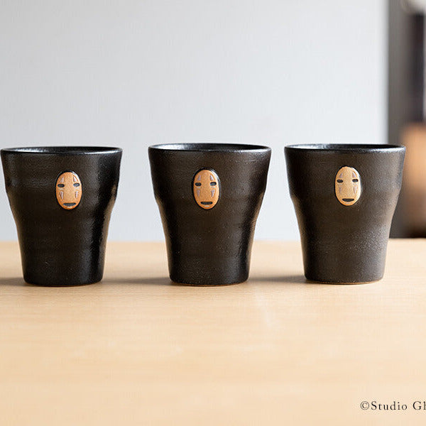 Three black ceramic cups with face designs on a wooden surface, featuring Studio Ghibli branding.