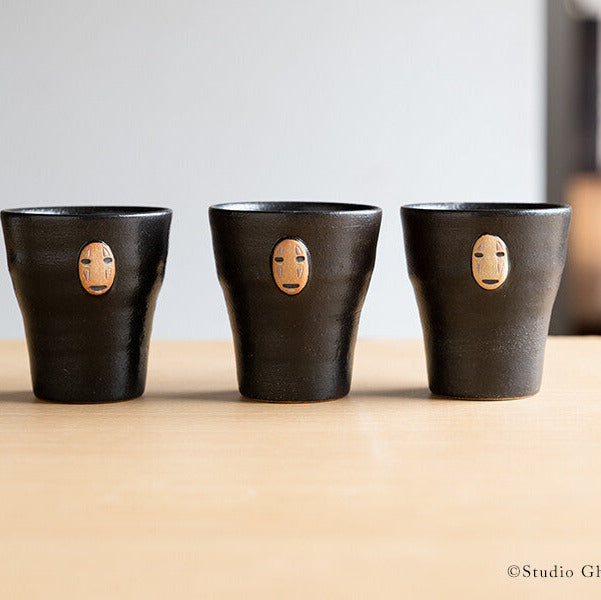 Three black ceramic cups with face designs on a wooden surface, featuring Studio Ghibli branding.
