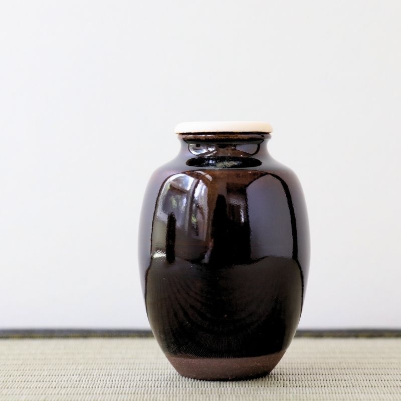 A glossy dark brown ceramic jar with a light beige lid, standing on a textured woven mat against a plain white background.