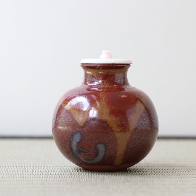 A small glossy ceramic lidded jar with a rounded body, featuring a mottled reddish-brown and amber-brown glaze with a dark gray abstract mark on the front, and a white lid, set against a neutral light gray background.