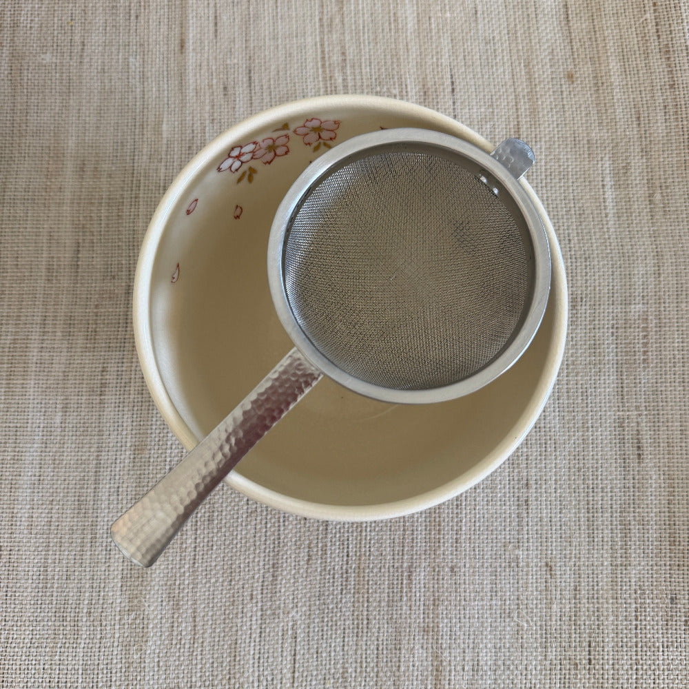 A stainless steel mesh sieve with a hammered metal handle rests inside a beige ceramic bowl decorated with a pink floral pattern, all placed on a textured beige woven placemat.