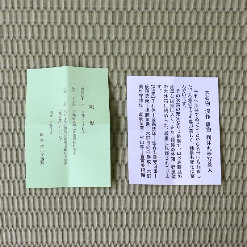 A folded light green booklet and a rectangular white card with Japanese text, placed on a woven straw mat background.