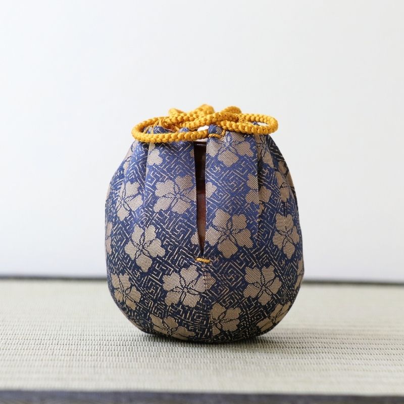 A small decorative fabric bag with a blue and beige floral pattern, featuring a drawstring closure at the top and a vertical slit, displayed on a textured surface against a plain light background.