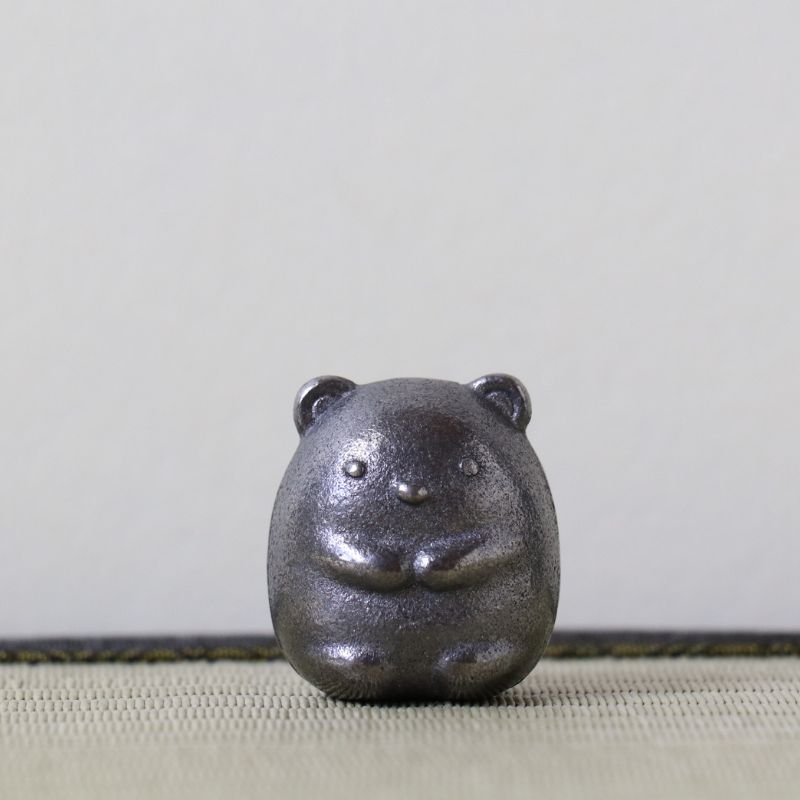 A small dark metallic bear figurine with a textured, hammered finish, sitting on a woven beige surface against a neutral light gray background.