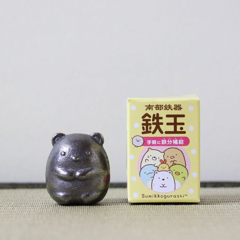 A small dark metallic Sumikkogurashi figurine stands next to its light yellow packaging box on a neutral surface against a light gray background.