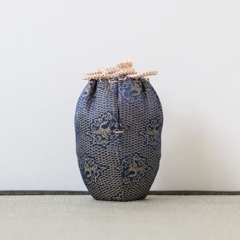 A small fabric drawstring pouch in dark blue and beige patterned material, standing upright against a plain light wall on a textured surface.