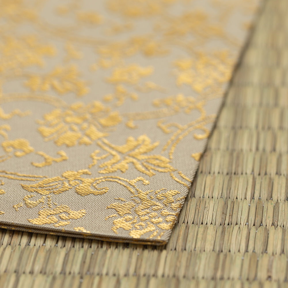 A close-up of a beige fabric or paper sheet with a metallic gold floral damask pattern, laid diagonally on top of a woven straw mat.