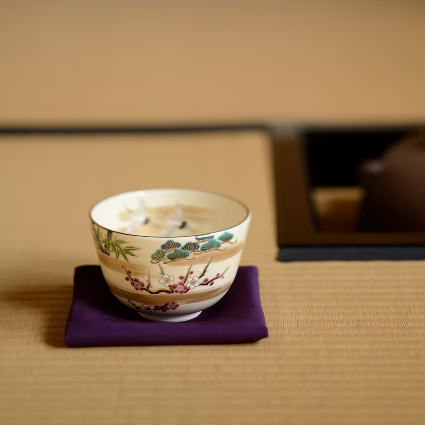 Crane and Sho-Chiku-Bai Kyo Ware matcha bowl with gold leaf accents displayed on a purple cloth in a Japanese tea room setting