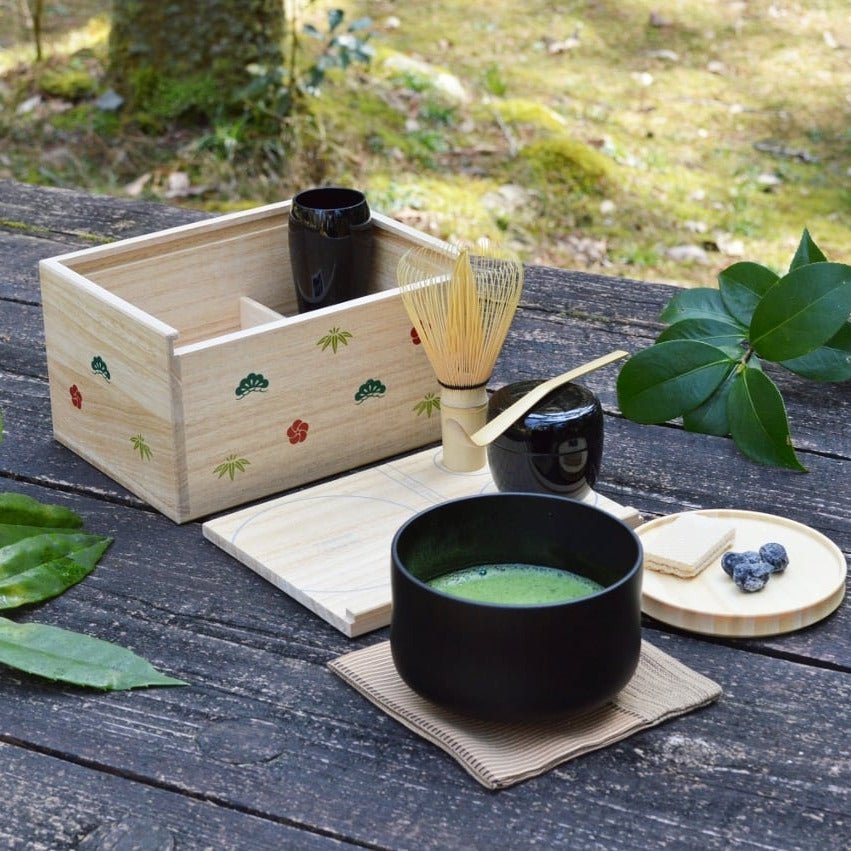 Tea-making setup with a black teapot, green tea, and leaves on a wooden table outdoors.