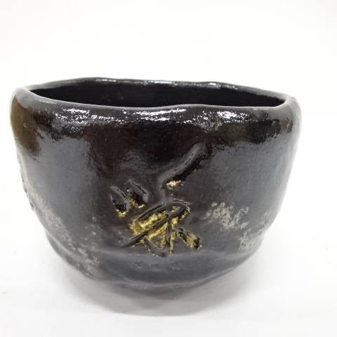 Front view of the Eiko Meido matcha bowl showing its softly irregular rim, smoky black glaze, pale markings, and gold botanical brushwork.