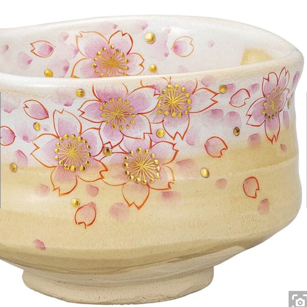 Left angled view of the Kutani Ware matcha bowl showing layered pink sakura blossoms, scattered petals, and gold details continuing around the exterior.