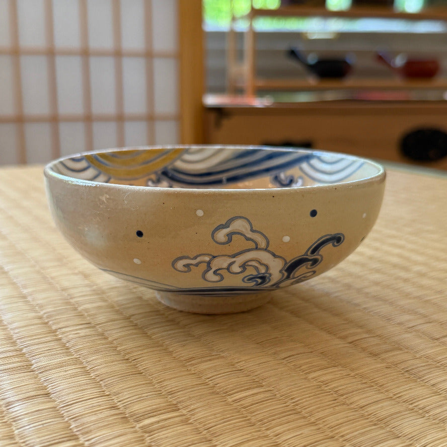 Front view of a hand-painted Kyo Ware matcha bowl with cobalt-blue curling waves, white foam, and ochre-gold accents on a cream glaze.