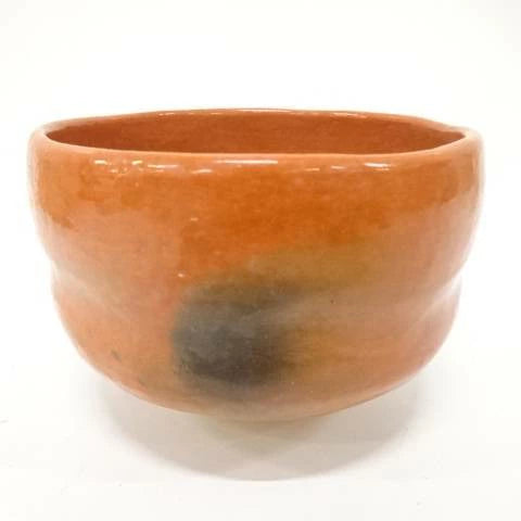 Front view of the Juraku Aka Raku chawan showing its warm coral-red glaze, rounded handcrafted form, and smoky black markings.