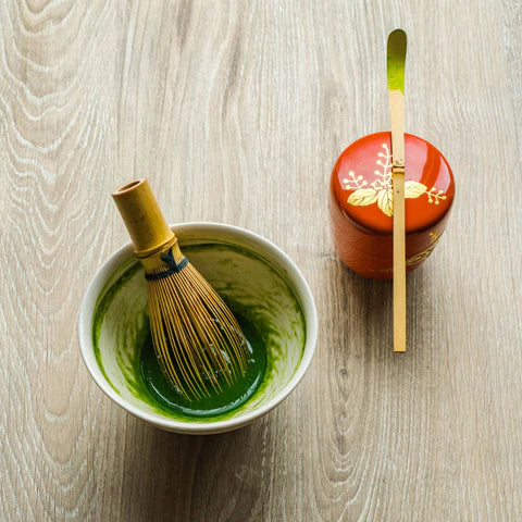 Bowl of green tea with a whisk and a small container on a wooden surface