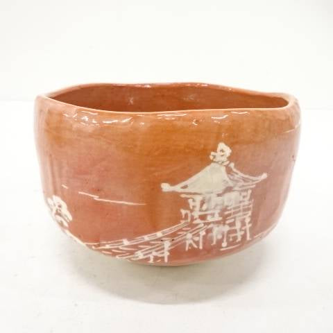 Front view of the Kawasaki Waraku Aka Raku chawan showing its coral-red glaze and white-painted pavilion, bridge, and garden foliage.