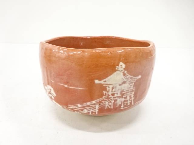 Front view of the Kawasaki Waraku Aka Raku chawan showing its coral-red glaze and white-painted pavilion, bridge, and garden foliage.