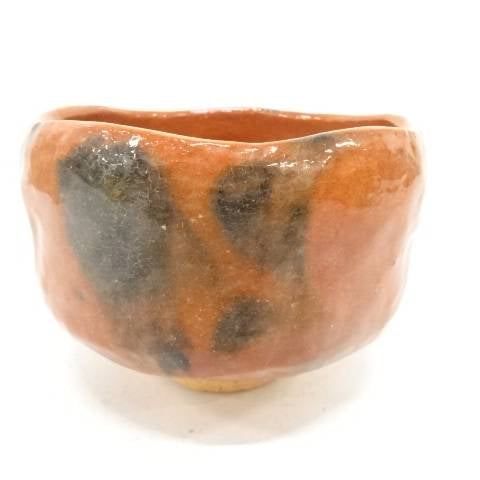 Front view of the Kawasaki Waraku Red Raku matcha bowl with warm red-orange glaze, charcoal-black markings, and a pale foot.