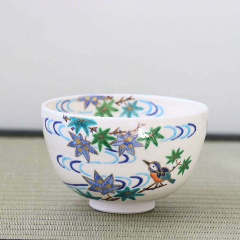 Angled view of the Kyo-yaki chawan showing a hand-painted bird, green maple leaves, and blue flowing water wrapping around the white glazed bowl