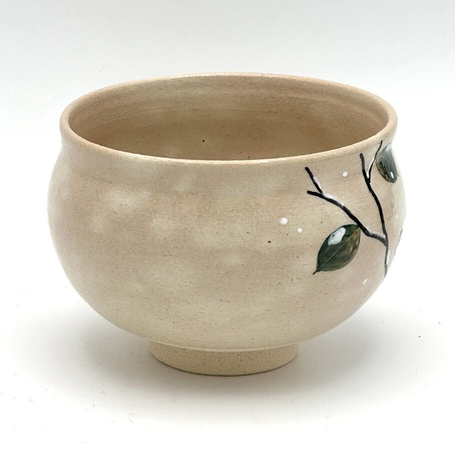 Side view of the hand-painted Kyo-yaki chawan showing a dark branch with green leaves and subtle white accents against the warm beige glaze
