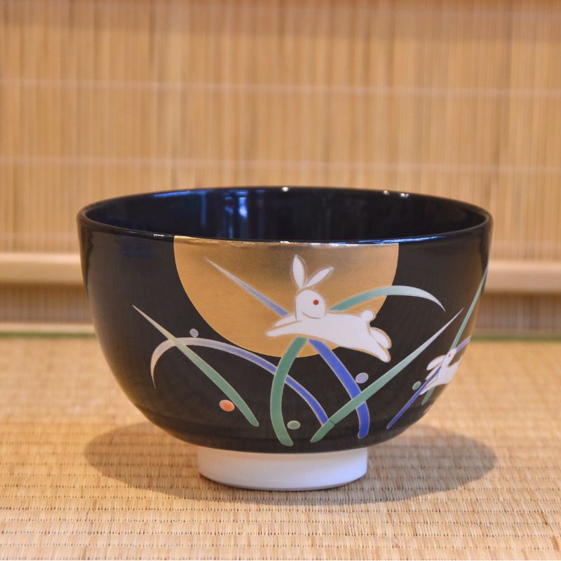 Front view of a black-glazed matcha bowl decorated with a white rabbit beneath a golden moon and colorful flowing grasses.