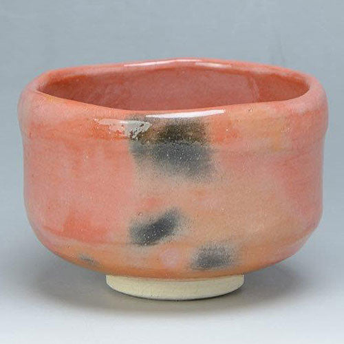 Front angled view of a handcrafted Aka Raku Rikyu chawan by Raku Zuisho with a coral-red glaze, smoky black accents, and a pale foot.