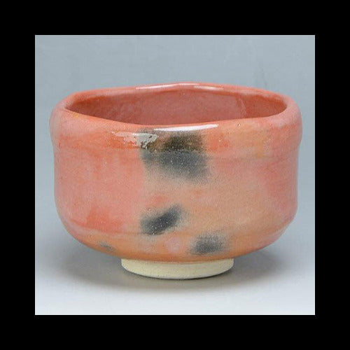 Front angled view of a handcrafted Aka Raku Rikyu chawan by Raku Zuisho with a coral-red glaze, smoky black accents, and a pale foot.