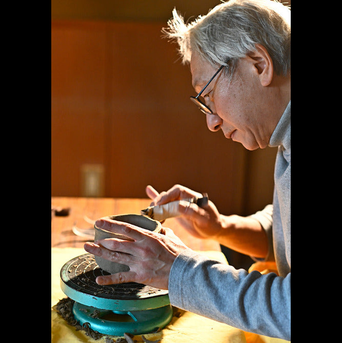An elderly potter with gray hair and glasses works at a pottery wheel, shaping a small clay vessel with a tool. The warm, golden light illuminates the potter's focused expression and the clay, set against a softly blurred brown background.