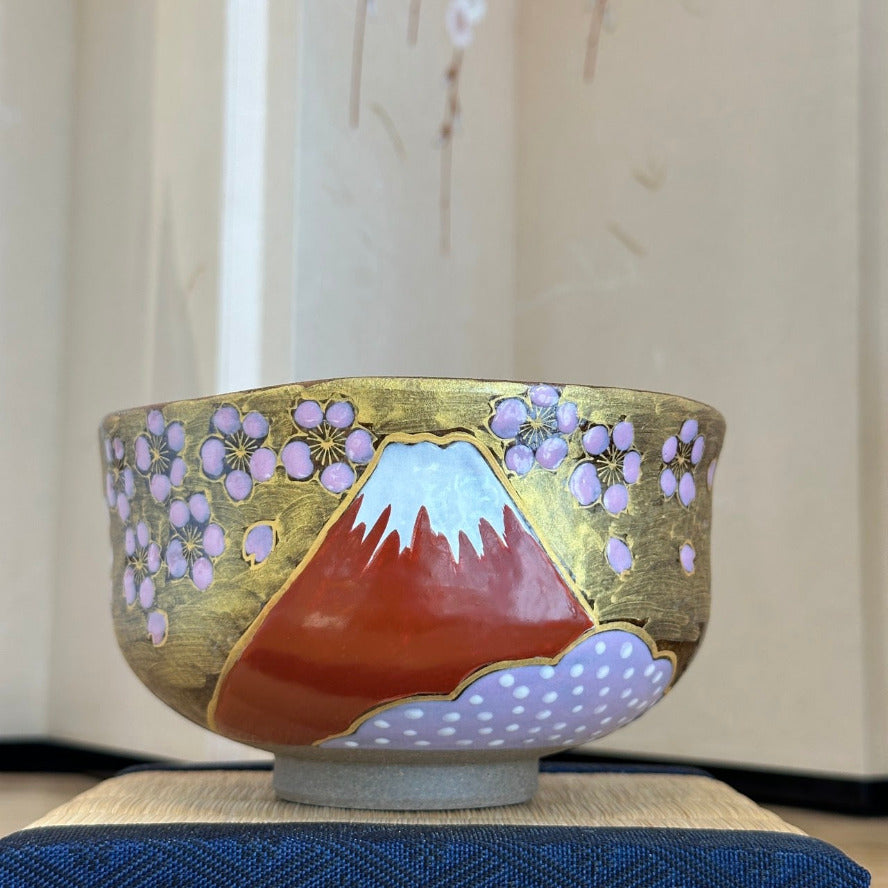 Front view of a hand-painted Kutani Ware matcha bowl featuring snowcapped Red Fuji, lavender sakura blossoms, and gold leaf, displayed on a dark blue cloth.