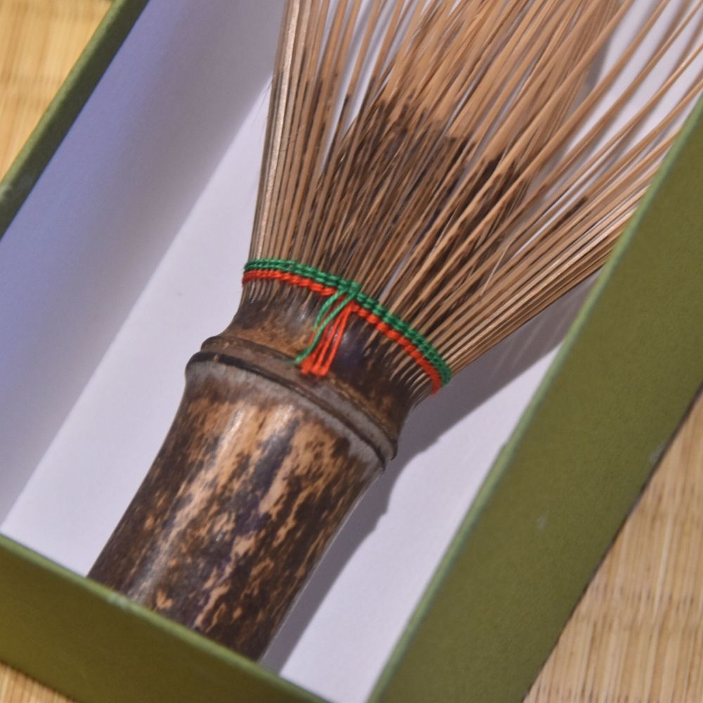 Traditional Japanese whisk with wooden black bamboo handle, cotton threads in red and green and thin bristles inside the green box with a light white inner background.