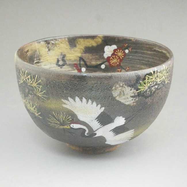 Front view of a hand-painted Sakata Yahata Kyo Ware matcha bowl with a large white crane, red and white plum blossoms, and gold-toned botanical accents on charcoal glaze.