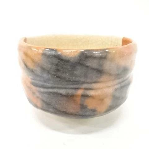 Side view of the Shoraku Sasaki Kotobuki Red Raku chawan showing its warm red-orange surface, hand-formed shape, and flowing dark charcoal glaze